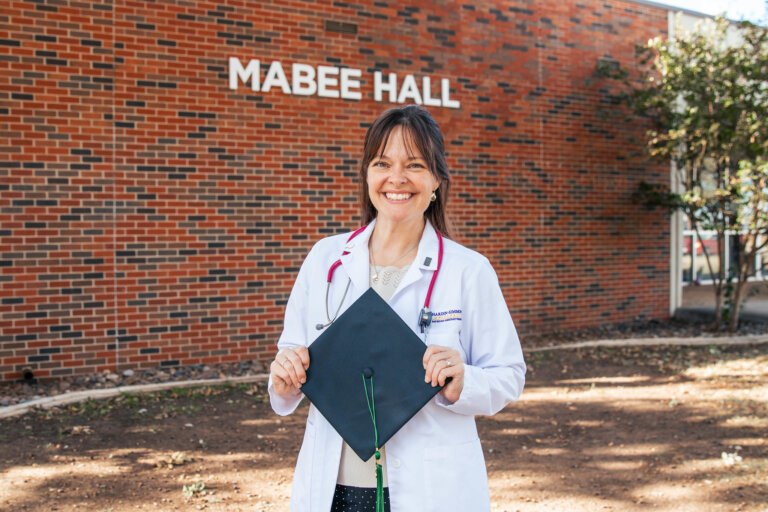 Bonnie Ressler stands outside Mabee Hall in her white coat after completing Hardin-Simmons University’s Physician Assistant program. The Air Force veteran will begin a neonatology fellowship in May, continuing her lifelong mission of caring for the vulnerable.